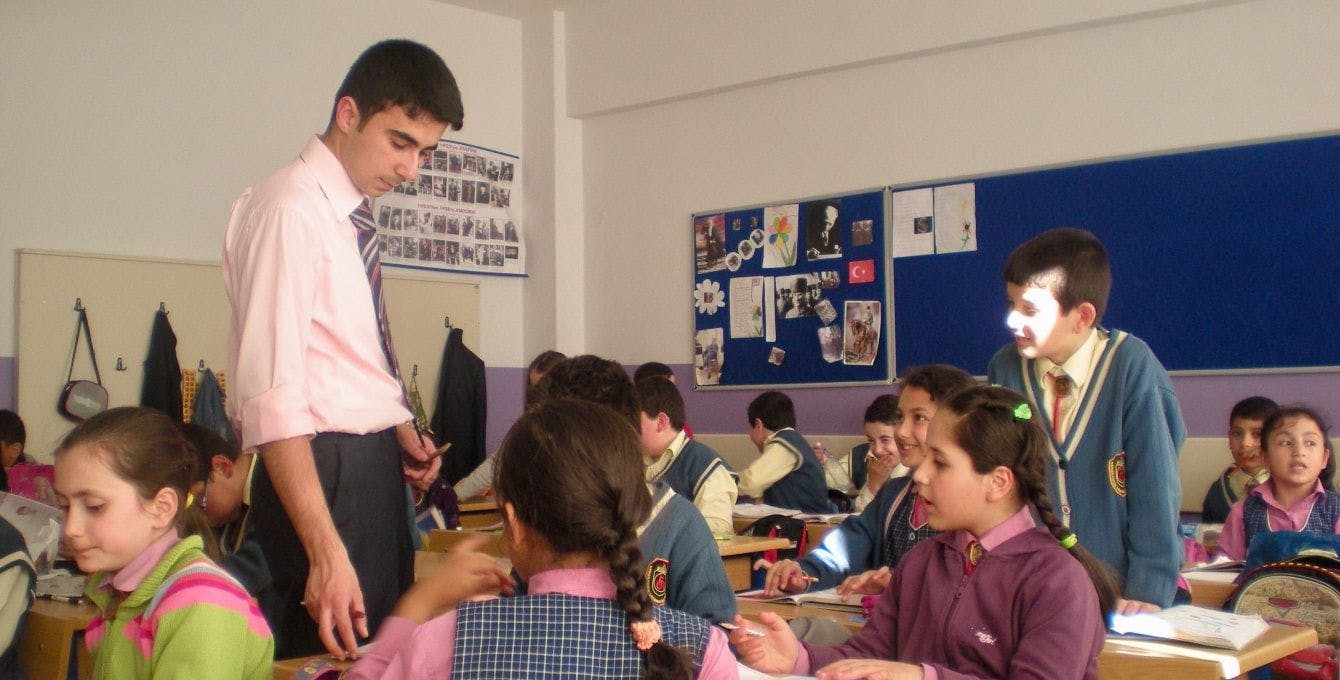 Menteş wears a suit and tie teaching students in a classroom.